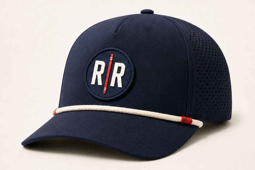 A navy blue baseball cap with a circular embroidered patch on the front featuring the letters 'R/R' in white with a red vertical line. A white rope detail with red end-caps is wrapped around the edge of the brim.